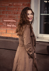 Fototapeta premium Young smiling woman walking on the street city wall background in brown coat and black shoes near the old bulding with big windows. Autumn