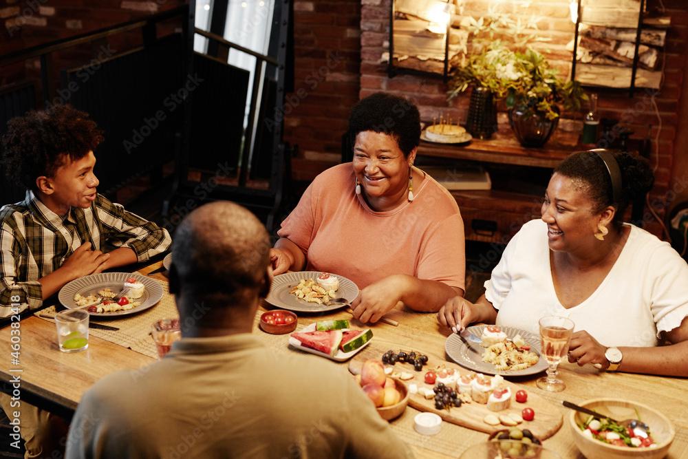 Wall mural Warm toned portrait of happy African-American family sitting at table while enjoying dinner together indoors at evening