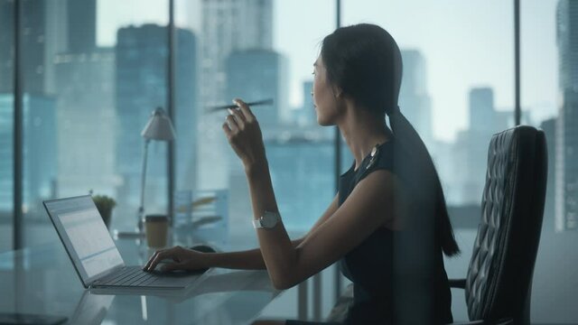 Successful Businesswoman in Stylish Dress Sitting at a Desk in Modern Office, Using Laptop Computer, Next to Window with Big City View. Successful Finance Manager Planning Work Projects.