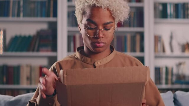 Unhappy young Afro American woman unpacking carton box feeling dissatisfied with getting wrong or damaged order, writing negative feedback in mobile application, internet shopping concept.