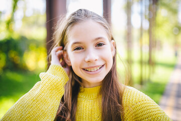 Cute and happy teen girl with braces smiling to camera in yellow clothes in park. Concept of dentist and orthodontist. Pretty teenage girl wearing teeth apparatus smile cheerfully outdoors