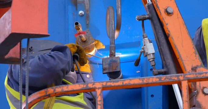 At the construction site welder in a vest and a protective helmet weld the part to a large blue color industrial equipment 