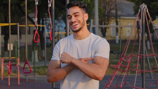 Pumped sportive millennial Middle Eastern Arab guy shows big biceps, proud of athletic achievements. Young Indian sportsman presents beautiful body muscles in morning workout outdoor at sports ground