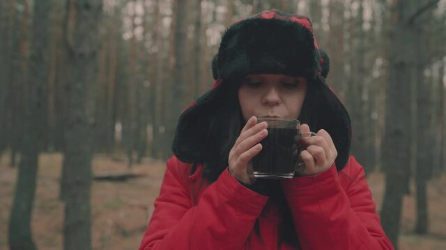 A portrait of the pretty woman with a cup of tea in woods. The woman in a jacket and a hat with earflaps is drinking a tea in a cold weather.