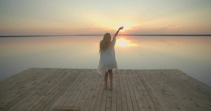 Woman with blonde hair is dancing on the pier of a lake during sunset, 4k