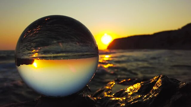 A glass ball stands in the foreground on a cliff in front of the sea with light waves at sunset in Kos, Greece