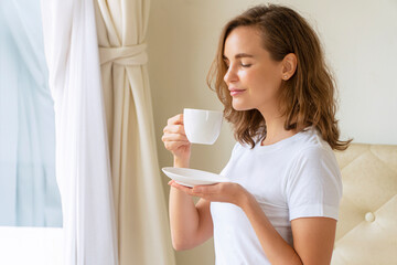 Young woman holding and drinking cup of coffee or hot chocolate after wake up in the morning while sitting on the bed feeling relaxed. Lifestyle concept.