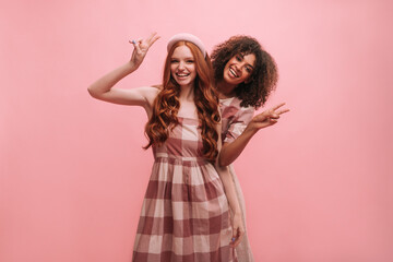 Medium shot of two interracial teenagers showing peace gesture with copy space. Happy curly-haired brunette hides behind long-haired red-haired girl. Female friendship concept