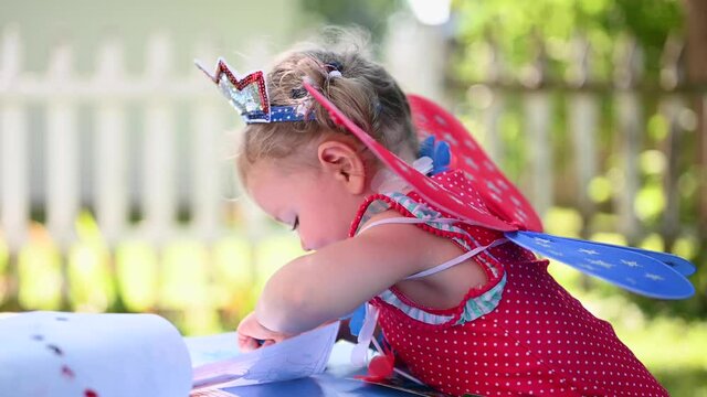 little girl in fairy wings and swimsuit drawing outside at family party on Fourth of July