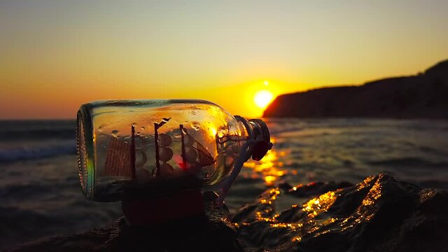 A ship in a bottle stands on a rock in front of a sunset while sea spray rushes up from the sea in slow motion, Kos, Greece - 2