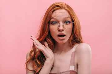 Close-up of surprised young fair-skinned red-haired girl on pink background. Portrait of astonished teenager with open mouth and transparent glasses. Concept of human emotions, facial expressions
