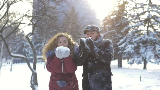 Slow motion. A beautiful romantic couple blows snowflakes from their palms on a bright sunny day in a snowy city park, cold winter along the Christmas trees. A tender relationship between lovers.