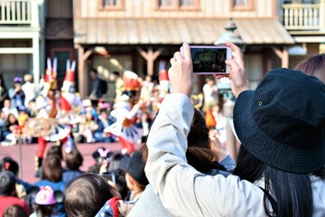A girl in a black bucket hat stands with the crowd and takes a photo of a parade with her phone 