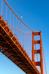 Closeup of portion of Golden Gate Bridge in San Francisco, California, with glowing crescent slivered moon and deep blue twilight sky.