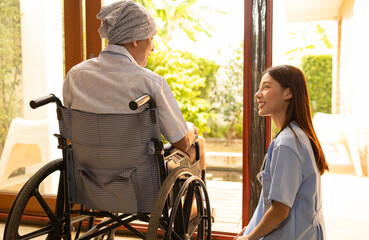 Attractive young Asian female nurse kneeling beside senior patient in wheelchair talking, smiling and cheering up in comfort at home.He survive from cancer .