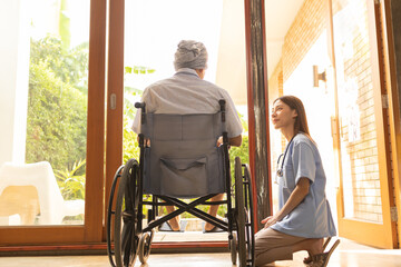 Attractive young Asian female nurse kneeling beside senior patient in wheelchair talking, smiling...
