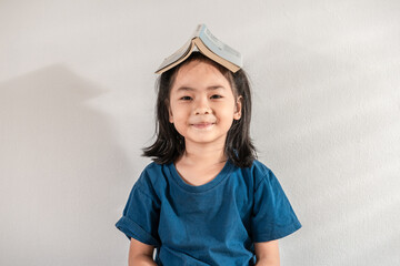 Portrait of a funny smile little toddler Asian student girl in a blue T-shirt with a book on her head at home school on white background. Education learning reading and child future success concept