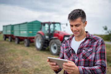 Portrait of happy farmer holding tablet standing in front of tractor with trailer.