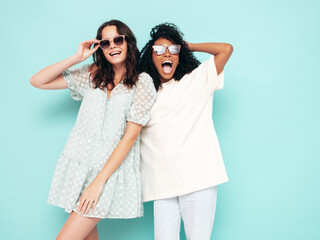 Two young beautiful smiling international hipster female in trendy summer clothes. Sexy carefree women posing near blue wall in studio. Positive models having fun in sunglasses. Concept of friendship