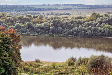 Panoramic view of a wide river with grassy banks and trees in the distance against grey clouds. Top view photo of early autumn in the countryside.