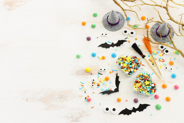 top view image of Halloween holoday. witcher hat, broom, bare trees, treats and bats over white wooden table