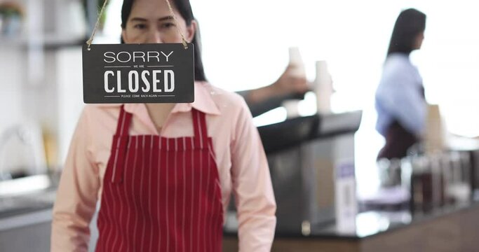 Cute Asian woman tie apron to fit body during walking to turn sign board at door of coffee shop from closed to open with cefe employees woking in blur background.