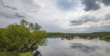 Landscape with a dramatic sky reflected in the river. Early spring, juicy May greens. Bright green foliage on trees and bushes.