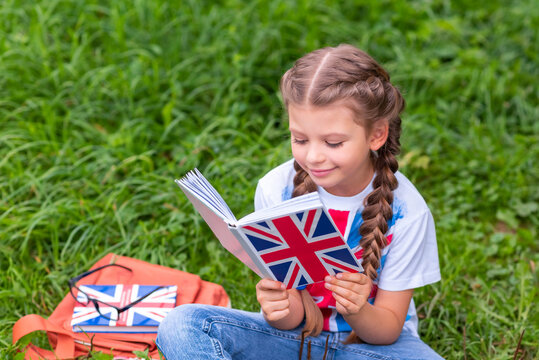 A little girl is studying a book on the English language sitting on the lawn.