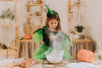 cute little girl in a green Halloween costume of a witch or fairy prepares a pumpkin pie, barm brack in a festive decorated kitchen. space for text. High quality photo