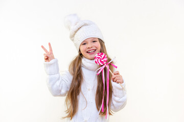 A little girl in a winter sweater and hat holds a delicious lollipop and smiles on a white isolated background.