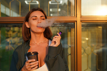 Woman with cup of coffee outdoor at city street at sunset with electronic cigarette catching sun...