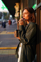 Woman with cup of coffee outdoor at city street at sunset with electronic cigarette catching sun rays enjoying smoking