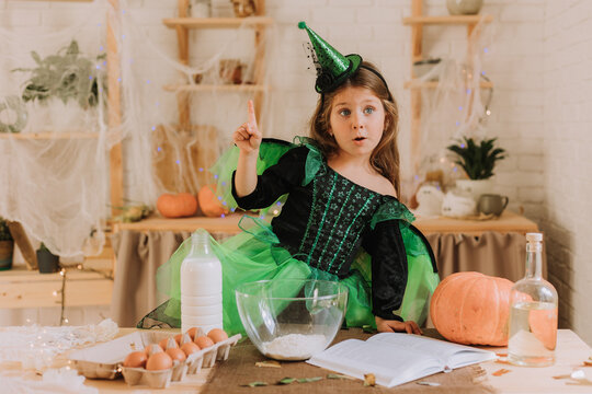 cute little girl in a green Halloween costume of a witch or fairy prepares a pumpkin pie, barm brack in a festive decorated kitchen. space for text. High quality photo