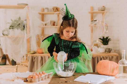 cute little girl in a green Halloween costume of a witch or fairy prepares a pumpkin pie, barm brack in a festive decorated kitchen. space for text. High quality photo