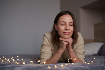 Pretty Caucasian woman smiling with fairy lights laying on bed. Comfy home. Dreaming time
