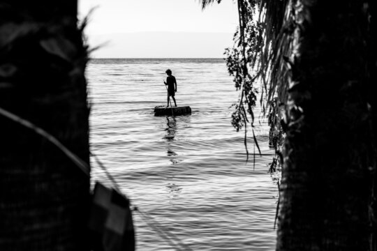 Black And White Photo Silhouette Of Adventurous Boy Sail On A Raft. Lake With Calm Water Between The Trees.