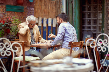 two men, young and senior, playing chess in outdoor cafe, smiling