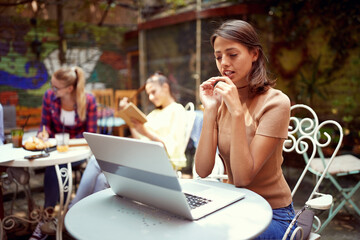 beautiful young woman sitting in outdoor cafe looking at her laptop on table, thinking