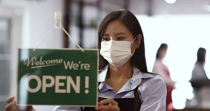 Cute Asian wears protective hygiene face mask hanging signboard at door of coffee shop with a happy face. Concept of reopening business after Covid-19 pandemic outbreak.  