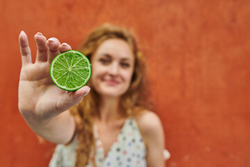 Red haired ukranian woman in casual dress playing with two halfs of lime on orange background