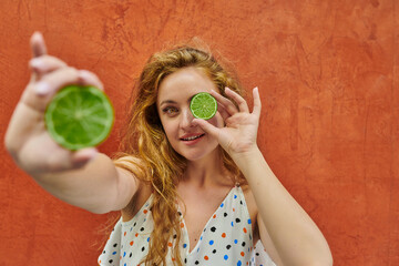 Red haired ukranian woman in casual dress playing with two halfs of lime on orange background