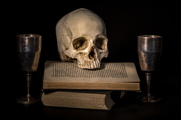 ludzka czaszka na książce i dwa metalowe kielichy na czarnym tle