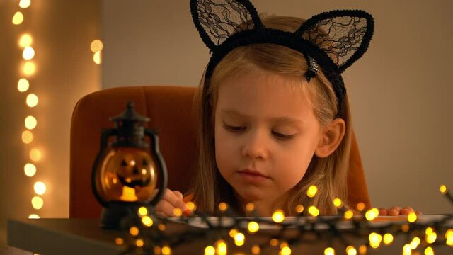 Child sits in dark with halloween pumpkin lamp and paints orange pencil on background of shining blurred lights of golden garland. Girl makes decoration for holiday