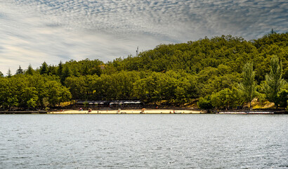 Cloudy sky over a sandy beach in Sanabria lake in Spain, surrounded by green vegetation, frequented by tourists all year round; view from the water.