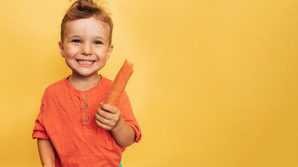 Studio shot of a smiling boy holding a fresh carrot on a yellow background. The concept of healthy...