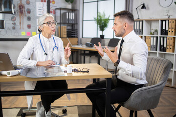 Aged female doctor sitting at own cabinet and doing regular check up for handsome caucasian businessman. Male patient visiting modern clinic for getting professional examination.