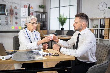 Caucasian senior woman in white labcoat and eyeglasses giving pills to male patient in formal wear at mededical cabinet. Concept of prescription and treatment.