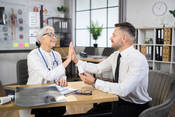 Happy man in business wear giving high five to female doctor at medical cabinet. Successful recovery after professional treatment. Medicine concept.