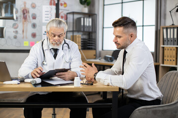 Senior bearded doctor in eyewear and lab coat writing on clipboard while listening to male patient in formal clothes. Caucasian businessman visiting competent therapist for health treatment.