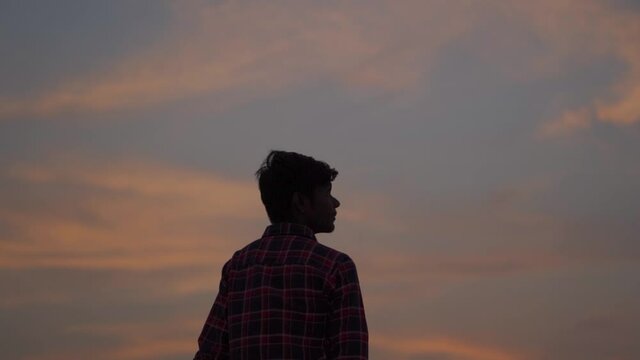 Silhouette shot of an Indian kid looking at the orange clouds during the sunset. Teenage kid looks at the sky in awe. Concept of hope and dreams in child. 